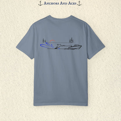 Blue Jean Comfort Colors 1717 water ski shirt shown from the back, featuring minimalist line art of a ski boat pulling a slalom water skier across calm lake water at sunrise with pine trees and blue spray.
